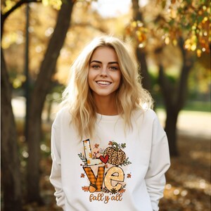 Op de afbeelding: Een witte sweatshirt met het woord "LOVE" in een herfstthema-ontwerp. Het ontwerp bevat een hart, een pompoen met luipaardprint en de woorden "fall y'all". De sweatshirt wordt gedragen door een persoon met blond haar.