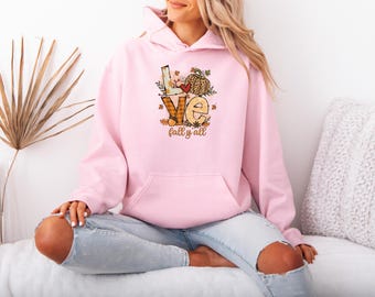 Könnte beinhalten: Ein hellrosa Kapuzenpullover mit einer Vordertasche. Das Design auf der Vorderseite zeigt das Wort "LOVE" in einer herbstlichen Schriftart, einen Kürbis und den Satz "fall y'all".