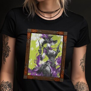 May include: Black t-shirt featuring a framed illustration of a cowgirl with two pistols, wearing a hat and purple accents. The artwork has a green and purple background, and the frame is brown.