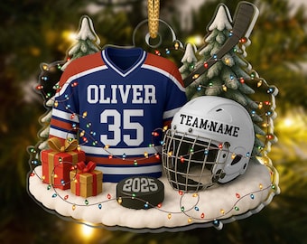 May include: A decorative ornament featuring a blue and orange hockey jersey with the name OLIVER and number 35, a white hockey helmet labeled TEAM-NAME, a black puck marked 2025, two gift boxes, and pine trees, all wrapped in colorful string lights.