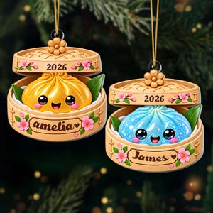 May include: Two hanging ornaments shaped like bamboo steamer baskets. One contains a golden yellow dumpling with the name amelia, the other a blue dumpling with the name James. Both feature the year 2026, pink cherry blossoms, and green leaves.