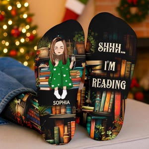 Op de afbeelding: Een paar zwarte sokken met een bedrukte illustratie van een persoon met bruin haar in een groene jurk met witte hartjes, lezend in een boek zittend tussen stapels boeken. De tekst luidt "SOPHIA" en "SHHH... I'M READING."