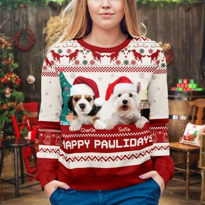 May include: A long-sleeve crewneck jumper featuring a red and white festive pattern with two dogs wearing Santa hats. The dogs are labelled Charlie and Sofia above the text HAPPY PAWLIDAYS. The design includes a Christmas tree and snowflakes.