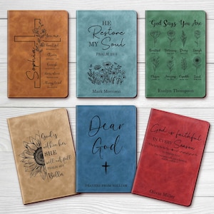 Op de afbeelding: Een verzameling gepersonaliseerde lederen journals in verschillende kleuren, waaronder bruin, blauw, groen en rood. Elk journal heeft unieke gegraveerde ontwerpen, zoals kruisen, bloemen en inspirerende citaten. Namen en zinnen zijn ook gegraveerd.