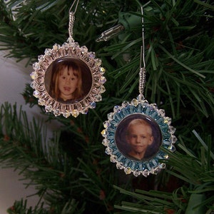 May include: Two Christmas ornaments with photos of children inside. The ornaments are made of clear glass beads with a silver wire hanger. The ornament on the left has a pink background and the ornament on the right has a blue background.