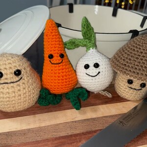 May include: A selection of crocheted vegetables with smiling faces, including a potato, carrot, onion, and mushroom. The handmade toys are arranged on a wooden chopping board near a large pot and a knife.
