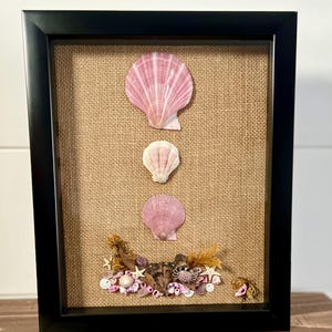 May include: A framed seashell art piece with a burlap background. Three pink seashells are arranged vertically in the center of the frame. The bottom of the frame is filled with smaller seashells and sea-themed decor.