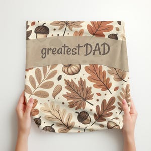 Op de afbeelding: Een beige deken met een herfstthema patroon van bladeren, eikels en pompoenen. De woorden "greatest DAD" zijn in bruin over het midden gedrukt. De deken wordt door twee handen omhoog gehouden.