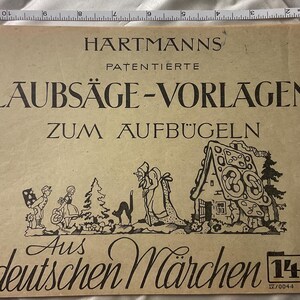 May include: A tan paper envelope titled HARTMANNS PATENTIERTE LAUBSÄGE-VORLAGEN ZUM AUFBÜGELN AUS DEUTSCHEN MÄRCHEN 141, featuring a black ink illustration of a fairy tale scene with a house, a cat, and figures, measured by a 12-inch ruler at the top.