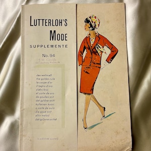 May include: Vintage fashion magazine cover featuring the text "LUTTERLOH'S MODE SUPPLEMENTE No. 94". The cover illustration depicts a woman in a red suit with a matching scarf and hat. The magazine is on a cream-colored surface.