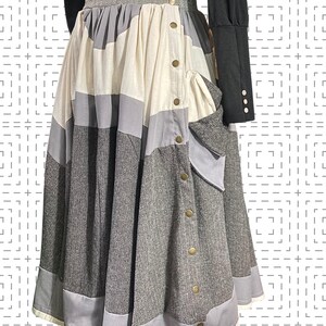 Op de afbeelding: Een patchwork rok in tinten grijs, crème en lichtblauw. De rok heeft een knoopsluiting aan de voorkant met goudkleurige knopen en een zak. De stof lijkt een mix van texturen te zijn, waaronder tweed en gladde materialen. De rok wordt gecombineerd met een zwarte top met lange mouwen.