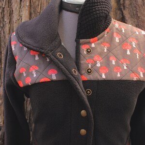 May include: A black fleece jacket with a brown and red mushroom print on the front. The jacket has a high collar and a zipper closure. It has a quilted design and a red trim around the pockets.