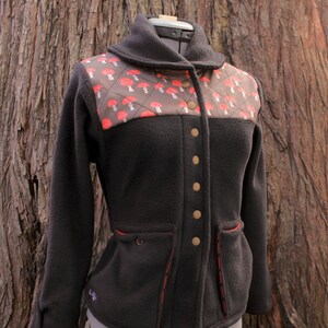 May include: A black fleece jacket with a red and brown mushroom print on the front. The jacket has a collar, a button closure, and two pockets.