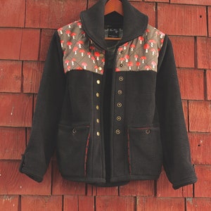 May include: A black fleece jacket with a mushroom print on the shoulders and a red stitching detail. The jacket has a zipper closure and multiple pockets.