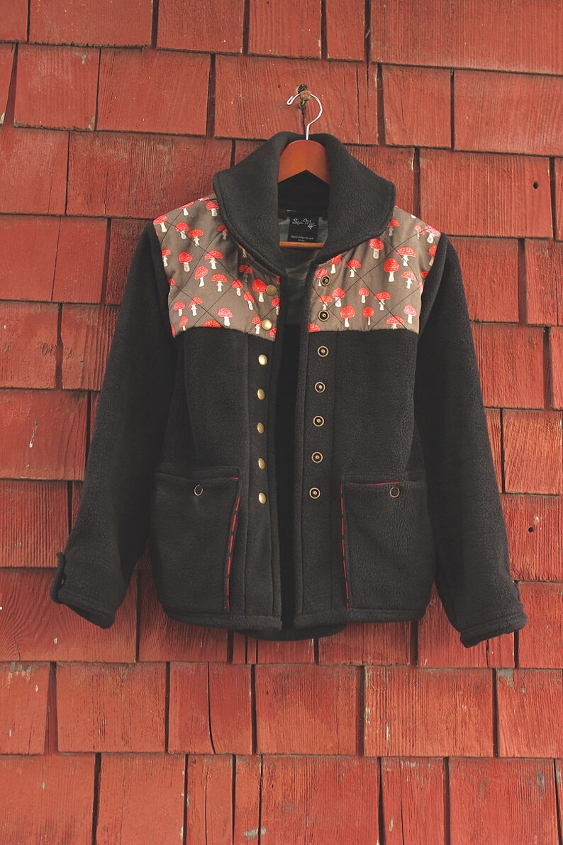 May include: A black fleece jacket with a mushroom print on the shoulders and a red stitching detail. The jacket has a zipper closure and multiple pockets.