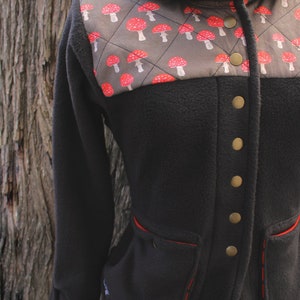 May include: A black fleece jacket with a brown and red mushroom print on the upper portion of the jacket. The jacket has a collar, a button closure, and red stitching around the pockets.
