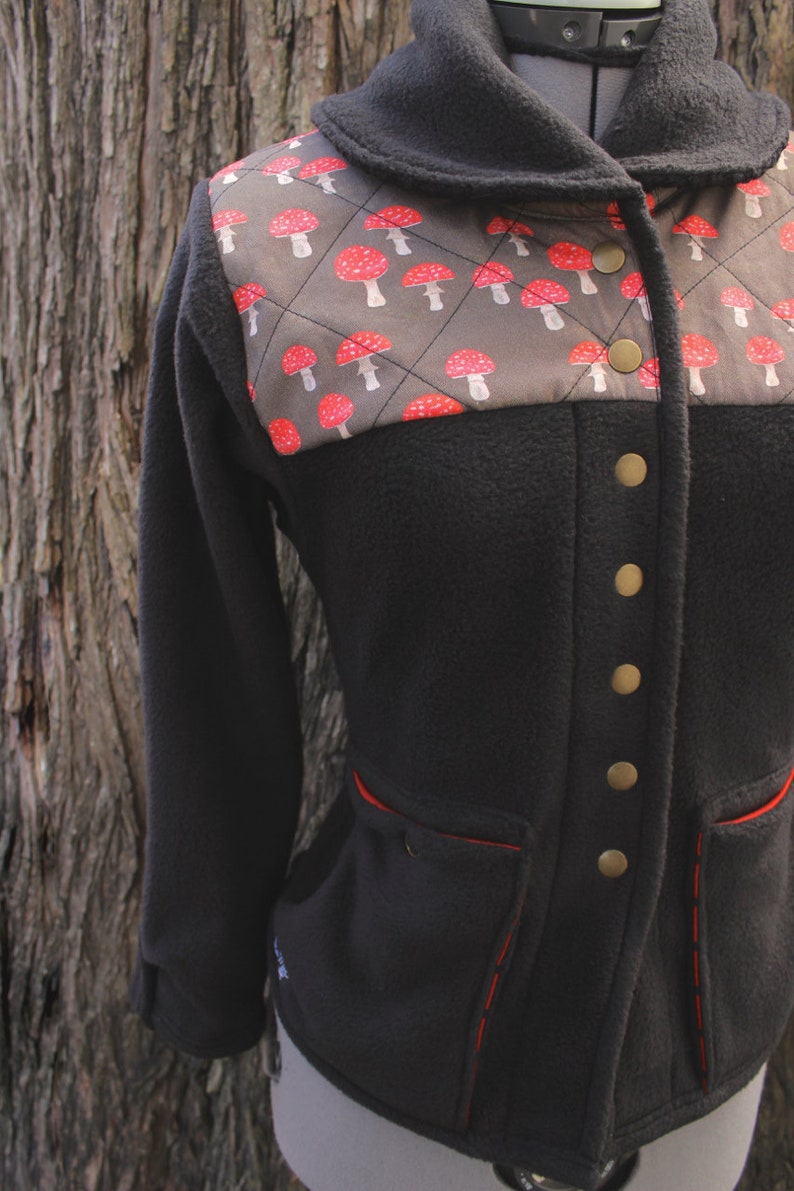 May include: A black fleece jacket with a brown and red mushroom print on the upper portion of the jacket. The jacket has a collar, a button closure, and red stitching around the pockets.