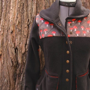 May include: Black fleece jacket with a brown and red mushroom pattern on the chest. The jacket has a high collar, a button closure, and two pockets with red stitching.