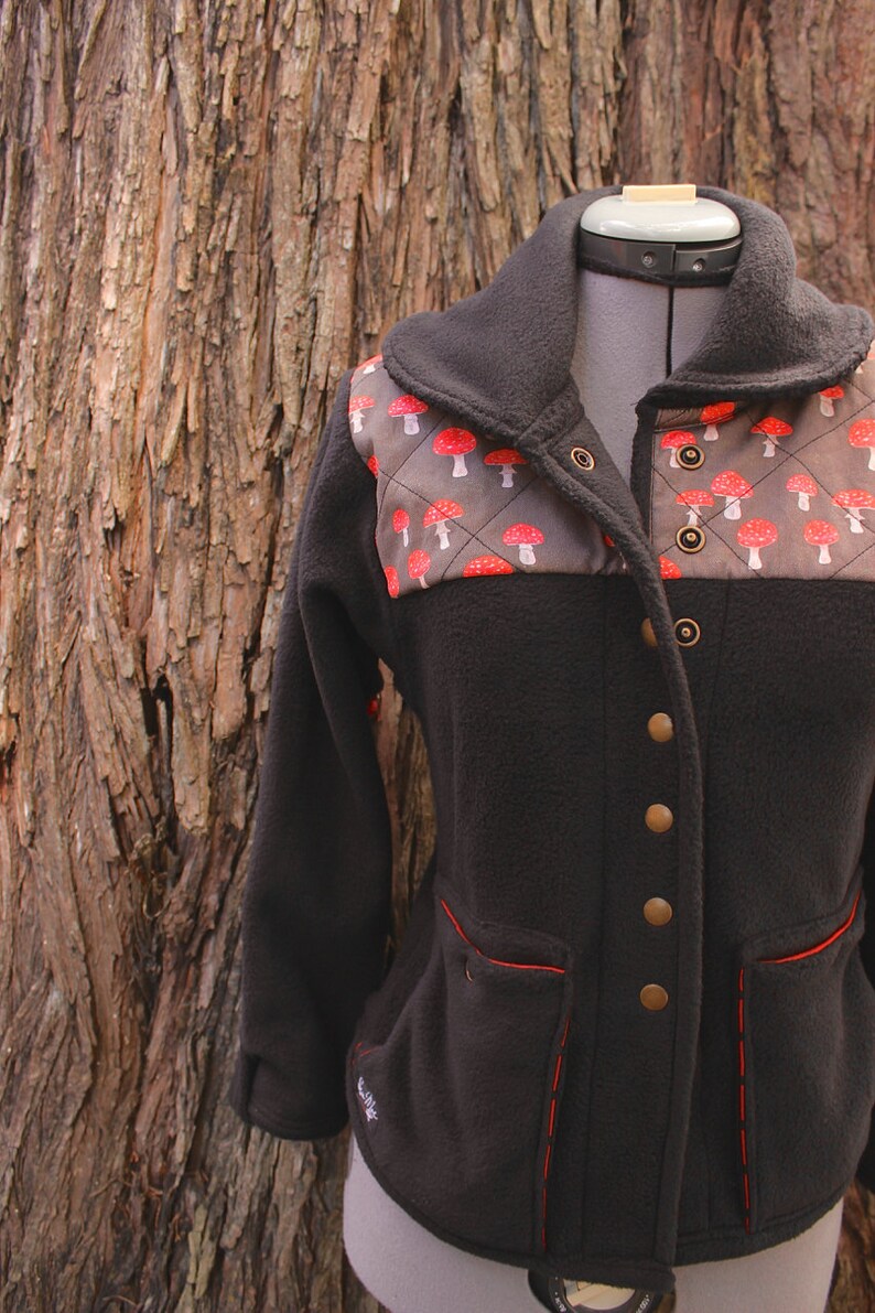 May include: Black fleece jacket with a brown and red mushroom pattern on the chest. The jacket has a high collar, a button closure, and two pockets with red stitching.