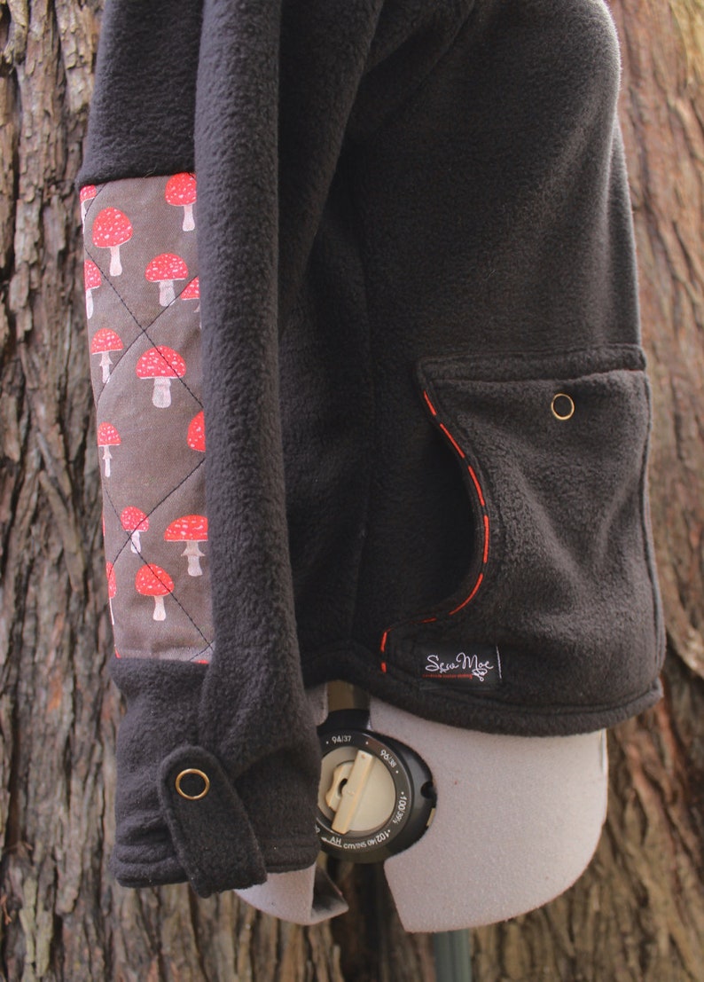 May include: Black fleece jacket with a red mushroom pattern on the left sleeve. The jacket has a red stitched pocket on the right side with the text "Sew Much" in white.