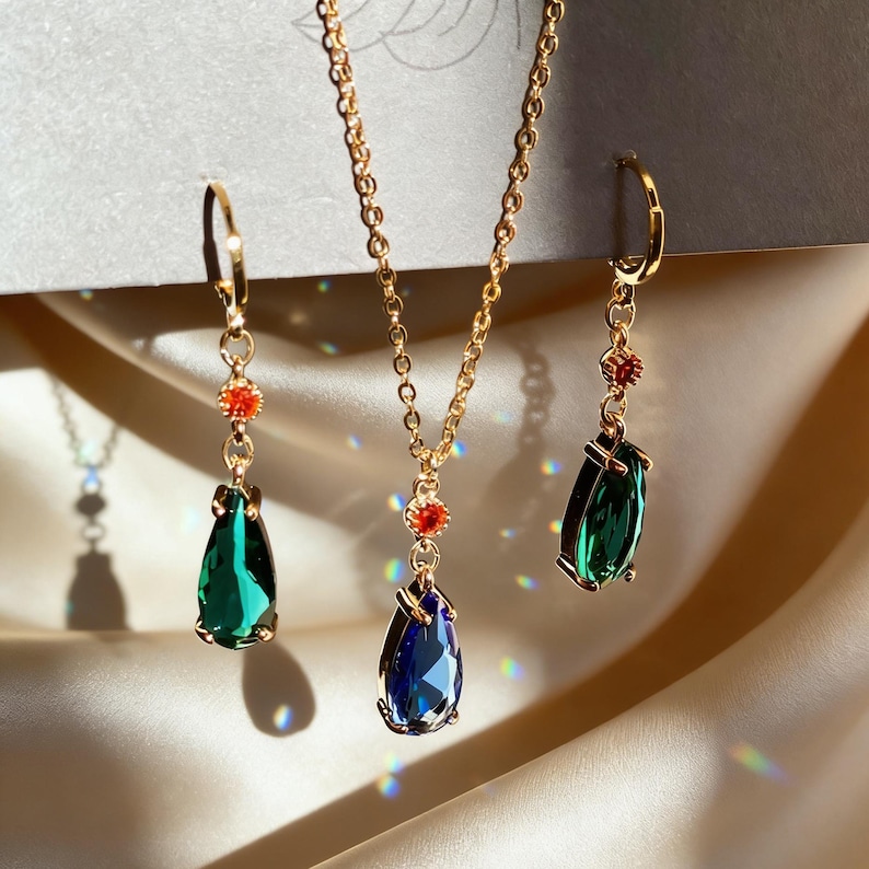 May include: A gold-toned jewelry set featuring a necklace and two earrings. The necklace has a blue teardrop-shaped pendant, while the earrings have green teardrop-shaped pendants. Each piece has a small red accent.