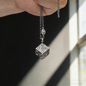 May include: A silver-toned necklace featuring a pendant shaped like a die. The die has six sides, each with eye-shaped markings. The necklace chain is also silver-toned and has a small eye-shaped charm near the clasp.
