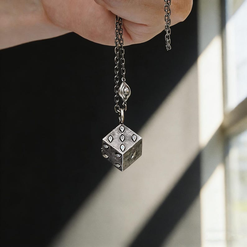 May include: A silver-toned necklace featuring a pendant shaped like a die. The die has six sides, each with eye-shaped markings. The necklace chain is also silver-toned and has a small eye-shaped charm near the clasp.