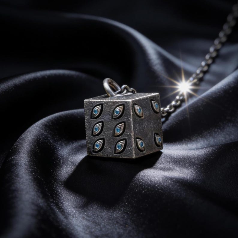 May include: A silver-toned cube pendant necklace with blue eye-shaped accents. The cube is suspended from a silver chain, resting on a black satin surface. The pendant has a textured appearance and a ring at the top for the chain.