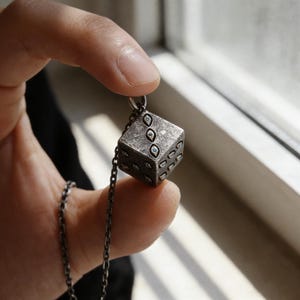 May include: A silver-toned pendant necklace featuring a cube-shaped charm designed to resemble a die. The die has small, round, blue accents in place of the dots. The necklace chain is also silver-toned.