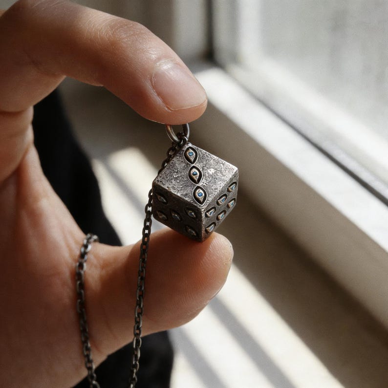 May include: A silver-toned pendant necklace featuring a cube-shaped charm designed to resemble a die. The die has small, round, blue accents in place of the dots. The necklace chain is also silver-toned.