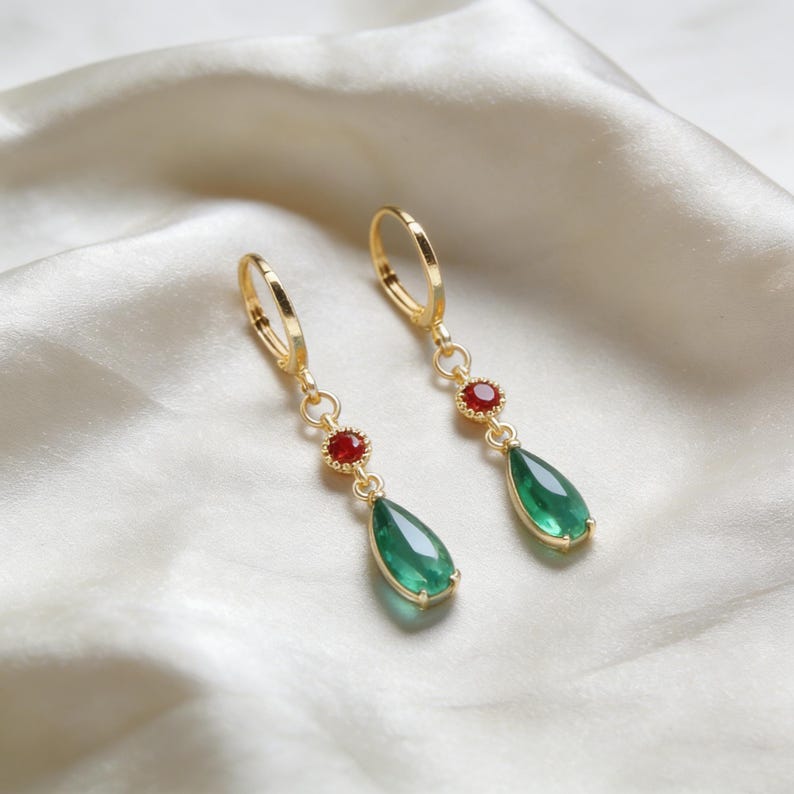 May include: Gold-tone earrings with a teardrop-shaped green gemstone and a small red gemstone. The earrings have a lever-back closure and are displayed on a white, silky surface. The earrings are a jewelry item.