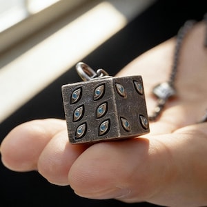 May include: A silver-colored pendant in the shape of a die, featuring eye-shaped designs with blue centers. The die is held in a hand, with a chain visible in the background. The pendant is a cube shape.