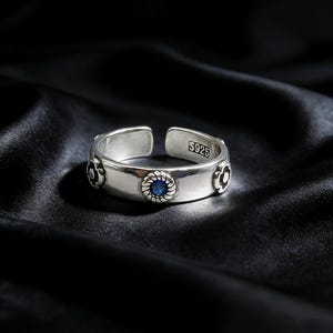 以下が含まれることがあります： 青い宝石を中央に、花のアクセントをあしらったシルバーの調節可能なリング。 リングには「S925」の刻印があり、宝石の周りにはロープのような縁取りがあります。 リングは黒いサテンの表面に表示されています。