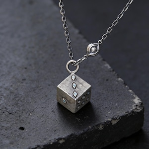 May include: A silver-toned necklace featuring a cube pendant with embedded blue gemstones. The cube has multiple sides with varying numbers of gemstone accents. The necklace chain is delicate, and the pendant is suspended from a small ring.