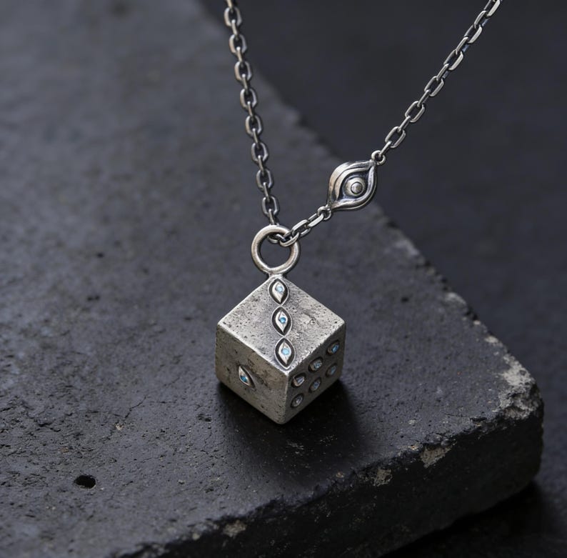 May include: A silver-toned necklace featuring a cube pendant with embedded blue gemstones. The cube has multiple sides with varying numbers of gemstone accents. The necklace chain is delicate, and the pendant is suspended from a small ring.