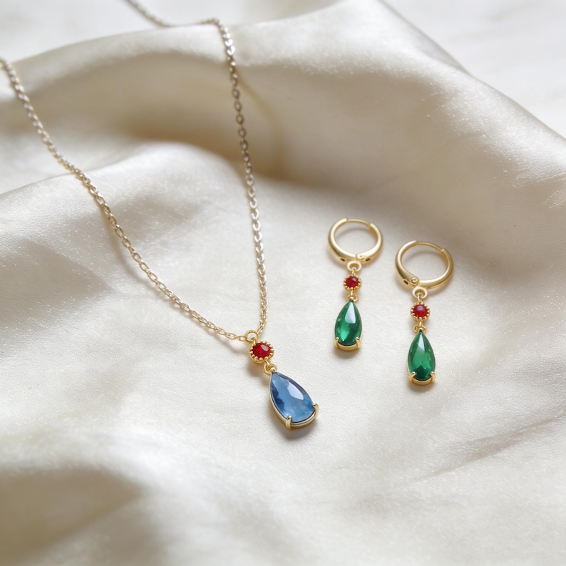 May include: A gold-colored necklace and matching earrings set. The necklace features a blue teardrop-shaped pendant with a red accent stone. The earrings have green teardrop-shaped pendants with red accent stones. The jewelry is displayed on a white satin surface.