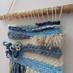 May include: A woven wall hanging with a driftwood rod at the top. The hanging features a variety of blue, white, and green yarn in a geometric pattern. The bottom of the hanging has long yarn strands in blue, white, and green.