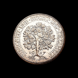 Puede incluir: Una moneda de plata con un diseño detallado de un árbol en el centro, rodeada por las palabras "EINIGKEIT UND RECHT UND FREIHEIT" y el año "1927". La moneda tiene un borde texturizado y está sobre un fondo negro.