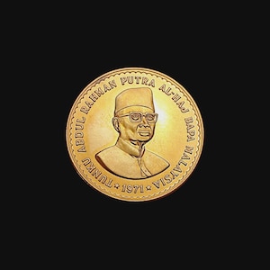 May include: A gold-colored coin featuring a portrait of a man wearing glasses and a hat. The coin has text around the edge that reads "ABDUL RAHMAN PUTRA AL-HAJ BAPA MALAYSIA TUNKU 1971". The coin is set against a black background.