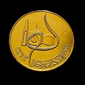 Könnte beinhalten: Eine goldfarbene Münze mit arabischer Kalligraphie und einem Bild der Kaaba, dem würfelförmigen Gebäude im Zentrum der heiligsten Moschee des Islam, der Masjid al-Haram in Mekka, Saudi-Arabien. Die Münze trägt auch den Text "15th C. H."