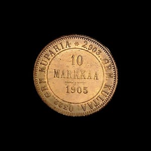 May include: A gold-colored coin with the text "10 Markkaa 1905" in the center. The coin's edge has a textured design, and the words "GRM KUPARIA" and "GRM KULTAA" are visible around the perimeter. The coin is set against a black background.