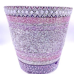 May include: A tapered cylindrical container covered in a dense layer of small, sparkling pink and silver beads. The top rim features a band of larger, diamond-shaped crystals arranged in a geometric pattern, while the base has a band of pink beads.