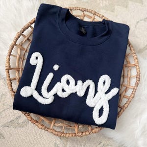 Może przedstawiać: Granatowa bluza z napisem "Lions" w białych, puszystych, haftowanych literach. Bluza prezentowana jest w wiklinowym koszu na dywanie w neutralnych odcieniach.