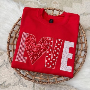 Può includere: Una felpa rossa con la scritta "LOVE" applicata sul davanti. La "L" è a quadretti rossi e bianchi, la "O" è un cuore con paillettes, la "V" è bianca con pois rossi e la "E" è con paillettes bianche. La felpa è esposta in un cesto intrecciato.