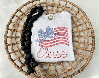 May include: White t-shirt with an embroidered American flag design featuring a blue bow with white stars and red and white stripes. The name "Eloise" is embroidered in red script below the flag. The shirt is displayed in a woven basket with black wooden beads.