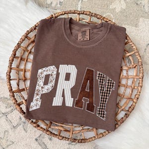 Könnte beinhalten: Ein braunes Comfort Colors T-Shirt mit dem Wort "PRAY" in einem Patchwork-Design. Die Buchstaben weisen florale, gestreifte, braune und karierte Muster auf. Das Shirt wird in einem Weidenkorb auf einer weißen und beigen Oberfläche präsentiert.