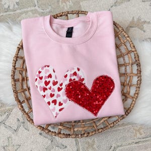 Pode incluir: Um moletom rosa claro com duas aplicações de coração. Um coração é coberto por corações vermelhos e rosa, e o outro é de lantejoulas vermelhas. O moletom está dobrado em uma cesta de vime.