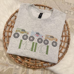 May include: A light gray sweatshirt with a crew neck, featuring an embroidered design of three monster trucks in green, blue, and tan, with the word "MAMA" in green below. The sweatshirt is folded in a woven basket.