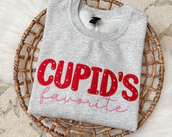 May include: A gray sweatshirt with the words "CUPID'S favorite" in red and pink embroidery. The red letters are decorated with small white hearts. The sweatshirt is displayed in a woven basket.