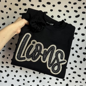 Könnte beinhalten: Schwarzes Sweatshirt mit dem Wort "Lions" in glitzernder Schreibschrift und goldener Umrandung. Der Ärmel ist sichtbar. Der Hintergrund ist weiß mit schwarzen Punkten. Das Sweatshirt ist gefaltet.